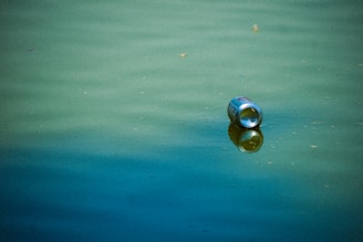 A metallic can floats on a body of greenish-blue water, casting a reflection on the surface. The water appears calm with slight ripples, and the can is the only visible object, indicating pollution or littering.