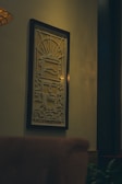 A framed artwork with intricate patterns hangs on a wall. The design features a symmetrical geometric pattern with a central figure resembling a stylized face, incorporating lines and shapes that create a sense of movement. The lighting casts a warm glow on the artwork and the surrounding space, which includes a partial view of a chair in the foreground and a hint of greenery at the bottom.