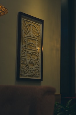 A framed artwork with intricate patterns hangs on a wall. The design features a symmetrical geometric pattern with a central figure resembling a stylized face, incorporating lines and shapes that create a sense of movement. The lighting casts a warm glow on the artwork and the surrounding space, which includes a partial view of a chair in the foreground and a hint of greenery at the bottom.