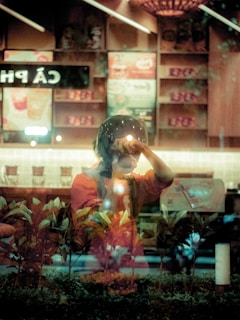 A person is seen through a reflective glass surface, holding a camera close to their face as if taking a picture. The setting appears to be an indoor cafe with menu signs visible in the background, partially obscured by reflections. The foreground displays a row of plants, adding a natural element to the scene.