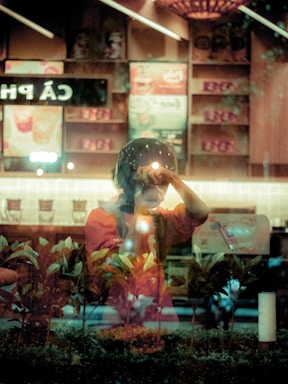 A person is seen through a reflective glass surface, holding a camera close to their face as if taking a picture. The setting appears to be an indoor cafe with menu signs visible in the background, partially obscured by reflections. The foreground displays a row of plants, adding a natural element to the scene.
