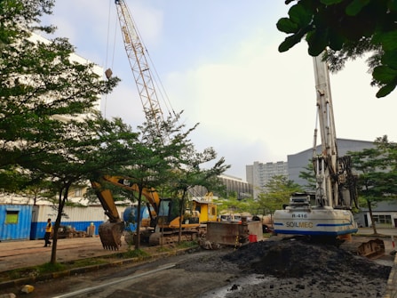 A construction site with heavy machinery, including a large crane and a Caterpillar excavator, surrounded by trees and dirt. Buildings are visible in the background with overcast skies above.