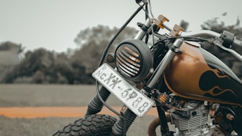 A close-up view of a motorcycle with a prominently displayed front number plate. The bike features a vintage headlight, intricate front suspension, and a custom paint job with flame graphics. The focus emphasizes the textures of the tire, metal, and paintwork against a blurred natural background.