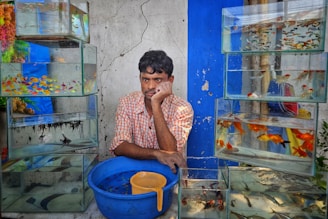 A man rests his head on his hand while sitting between stacks of glass aquariums filled with various colorful fish. He wears a checked shirt and is positioned in front of a weathered, cracked wall with blue accents. A large blue bucket with a yellow scoop is in front of him.