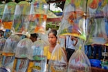 A market scene with clear plastic bags hanging from a wooden frame, each containing water and colorful live fish. A young woman stands behind the bags, wearing a yellow polka-dotted shirt, looking intently to the side. In the background, there are additional people who seem to be engaged in market activities. Trees and a structure with greenery on top are visible above.