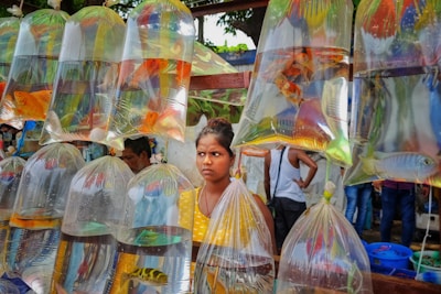 A market scene with clear plastic bags hanging from a wooden frame, each containing water and colorful live fish. A young woman stands behind the bags, wearing a yellow polka-dotted shirt, looking intently to the side. In the background, there are additional people who seem to be engaged in market activities. Trees and a structure with greenery on top are visible above.