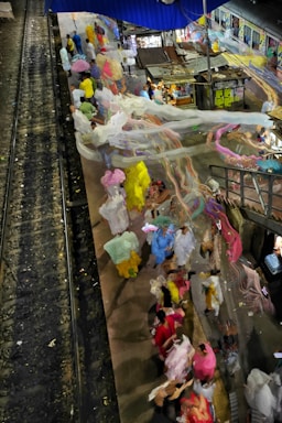A busy train platform with numerous people wearing colorful clothes and carrying bags. Motion blur creates a dynamic, flowing effect, emphasizing the hustle and bustle. The platform is adjacent to railway tracks, with a train partially visible at the edge of the image. Situated under a blue canopy, various small shops and stands can be seen.
