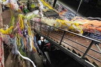 A vibrant and dynamic scene featuring a blurred, distorted view of a market or crowded area. The image includes a variety of colors with swirling patterns and motion blur, suggesting activity and movement. Metal railings and structures are visible, possibly indicating an overpass or bridge.