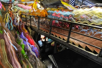 A narrow metal footbridge crosses between two levels, with the mesh floor casting shadows. On one side, vibrant, abstract streaks of multicolored fabric-like textures create a dynamic and chaotic effect. Below the bridge, an assortment of objects with bright colors is visible, suggesting a busy, crowded space.