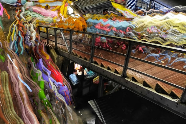 A narrow metal footbridge crosses between two levels, with the mesh floor casting shadows. On one side, vibrant, abstract streaks of multicolored fabric-like textures create a dynamic and chaotic effect. Below the bridge, an assortment of objects with bright colors is visible, suggesting a busy, crowded space.