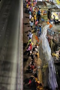 A crowded train station platform is bustling with people, many wearing vibrant clothing. The scene captures the motion and energy of the crowd, with a train passing by and market stalls visible in the background.