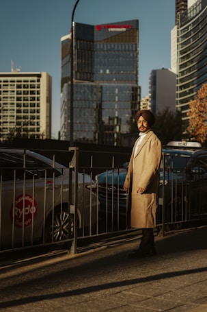 A man wearing a long beige coat and a turban stands on a sidewalk beside a row of cars. The background features modern, glass-covered buildings reflecting the late afternoon sun, casting warm shadows. A taxi sign is partially visible on one of the cars.