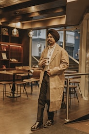 A man wearing a turban and a beige overcoat holds a coffee cup while standing in a modern cafe with wooden decor. Pendant lights hang from the ceiling, and there are tables and chairs in the background.