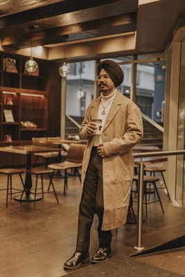 A man wearing a turban and a beige overcoat holds a coffee cup while standing in a modern cafe with wooden decor. Pendant lights hang from the ceiling, and there are tables and chairs in the background.
