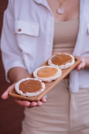 A person holds a wooden tray with three rice cakes spread with a brown topping. The individual is dressed in a light-colored outfit, wearing a pendant necklace. The background is blurred, emphasizing the focus on the rice cakes.