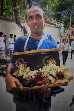 A person is holding a decorative tray adorned with floral garlands and ornate objects resembling traditional Indian headpieces. The background includes other people in casual attire and trees along a street.