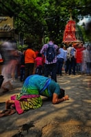A person in a colorful sari is bowing down on the street in a gesture of reverence or prayer. In the background, a crowd of people is gathered around a large, ornate red structure, possibly part of a religious festival or procession. The surrounding area is shaded by lush green trees, creating a vibrant and dynamic scene.
