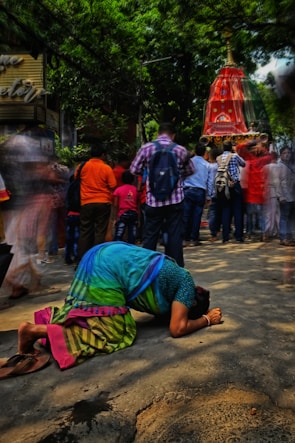 A person in a colorful sari is bowing down on the street in a gesture of reverence or prayer. In the background, a crowd of people is gathered around a large, ornate red structure, possibly part of a religious festival or procession. The surrounding area is shaded by lush green trees, creating a vibrant and dynamic scene.
