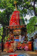 A vibrant and intricately decorated chariot is surrounded by people dressed in traditional attire. The chariot features ornate patterns with bright colors, including red and green fabrics, and is adorned with garlands and symbolic motifs. Lush green trees form a canopy above, and the ground is embellished with colorful rangoli patterns.