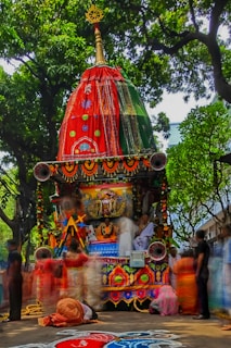 A vibrant and intricately decorated chariot is surrounded by people dressed in traditional attire. The chariot features ornate patterns with bright colors, including red and green fabrics, and is adorned with garlands and symbolic motifs. Lush green trees form a canopy above, and the ground is embellished with colorful rangoli patterns.