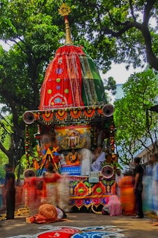 A vibrant and intricately decorated chariot is surrounded by people dressed in traditional attire. The chariot features ornate patterns with bright colors, including red and green fabrics, and is adorned with garlands and symbolic motifs. Lush green trees form a canopy above, and the ground is embellished with colorful rangoli patterns.