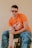 A person with short, dark curly hair is wearing large, rectangular glasses. They are dressed in an orange T-shirt with a race car graphic and the number 20, paired with blue jeans. The person is sitting on a black chair against a plain beige background, with one arm resting on their thigh.