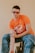 A person with short, dark curly hair is wearing large, rectangular glasses. They are dressed in an orange T-shirt with a race car graphic and the number 20, paired with blue jeans. The person is sitting on a black chair against a plain beige background, with one arm resting on their thigh.