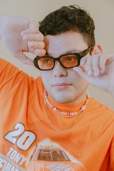 A person wears a bright orange T-shirt with graphic designs, layered necklaces, and black sunglasses with tinted lenses while holding the glasses with both hands. The background is a neutral tone.