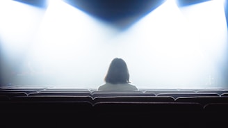 A person with shoulder-length hair sits alone in a dimly lit theater, facing the front. The bright stage lights create a high-contrast scene, casting the rest of the room into shadow.