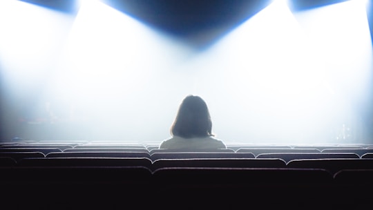 A person with shoulder-length hair sits alone in a dimly lit theater, facing the front. The bright stage lights create a high-contrast scene, casting the rest of the room into shadow.