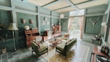 A vintage-style living room with teal walls and a high ceiling. The room features a collection of mid-century modern furniture, including a green sofa, armchairs, and a wooden coffee table. A wooden sideboard is adorned with globes and books, and a vintage television adds to the retro charm. Decorative elements include mounted deer antlers, a standing lamp, potted plants, and patterned curtains allowing natural light. The floor is covered with floral and geometric patterned tiles.