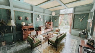 A vintage-style living room with teal walls and a high ceiling. The room features a collection of mid-century modern furniture, including a green sofa, armchairs, and a wooden coffee table. A wooden sideboard is adorned with globes and books, and a vintage television adds to the retro charm. Decorative elements include mounted deer antlers, a standing lamp, potted plants, and patterned curtains allowing natural light. The floor is covered with floral and geometric patterned tiles.