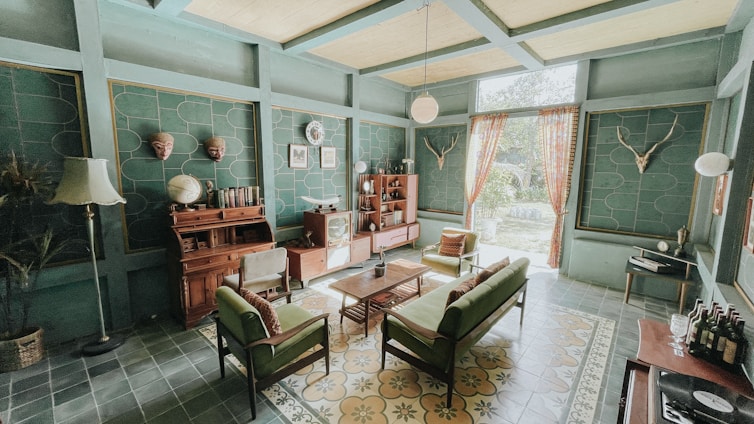 A vintage-style living room with teal walls and a high ceiling. The room features a collection of mid-century modern furniture, including a green sofa, armchairs, and a wooden coffee table. A wooden sideboard is adorned with globes and books, and a vintage television adds to the retro charm. Decorative elements include mounted deer antlers, a standing lamp, potted plants, and patterned curtains allowing natural light. The floor is covered with floral and geometric patterned tiles.