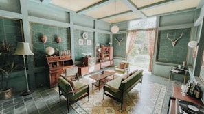 A vintage-style living room with teal walls and a high ceiling. The room features a collection of mid-century modern furniture, including a green sofa, armchairs, and a wooden coffee table. A wooden sideboard is adorned with globes and books, and a vintage television adds to the retro charm. Decorative elements include mounted deer antlers, a standing lamp, potted plants, and patterned curtains allowing natural light. The floor is covered with floral and geometric patterned tiles.