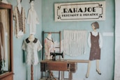 A vintage sewing studio with a wooden sewing machine table positioned against a light blue wall. On the wall, a sign reads 'RAHAJOE PENJAHIT & PENATOE' and features decorative elements. A macramé wall hanging adds texture beside a painted figure of a woman in a dress. Two dress forms stand nearby, each displaying different fabric garments.