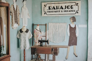 A vintage sewing studio with a wooden sewing machine table positioned against a light blue wall. On the wall, a sign reads 'RAHAJOE PENJAHIT & PENATOE' and features decorative elements. A macramé wall hanging adds texture beside a painted figure of a woman in a dress. Two dress forms stand nearby, each displaying different fabric garments.