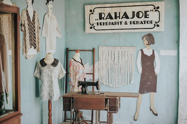 A vintage sewing studio with a wooden sewing machine table positioned against a light blue wall. On the wall, a sign reads 'RAHAJOE PENJAHIT & PENATOE' and features decorative elements. A macramé wall hanging adds texture beside a painted figure of a woman in a dress. Two dress forms stand nearby, each displaying different fabric garments.