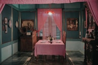 A cozy dining room with vintage decor, featuring a table covered with a pink tablecloth set for a meal with wine glasses and a candle holder. The walls are adorned with ornate frames and decorative plates. A gramophone sits atop a wooden cabinet, next to a vase with flowers. Pink curtains frame a window, and a chandelier with a red shade casts a warm glow.