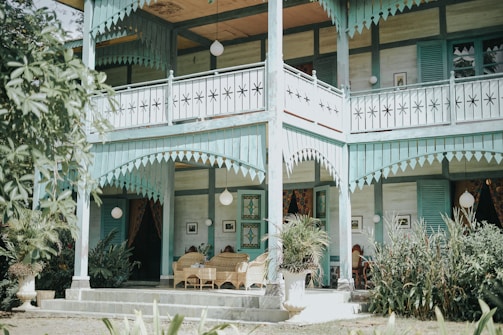 A charming two-story wooden house with decorative elements and pastel colors. The exterior features intricate woodwork on the balcony and verandah, and there are potted plants adding greenery around the entrance. Wicker chairs are placed on the porch, providing a cozy and inviting atmosphere. Shuttered windows and hanging lanterns enhance the vintage look.