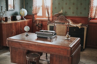 A vintage office setting featuring a dark wooden desk with a typewriter on it. There are two globes on the desk and a small wooden stool nearby. The room has wooden furniture, including a sideboard with decorative items, and walls adorned with patterned wallpaper. Light drapes partially cover the windows, and a mounted antler decorates the wall.