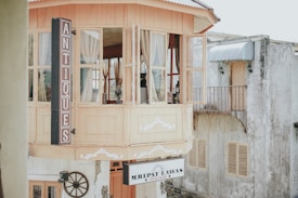 A charming antique shop with a rustic wooden facade, featuring large windows framed with cream-colored curtains. Above the entrance is a vertical sign reading 'ANTIQUES'. Below, a round decorative piece and a sign saying 'MRIPAT LAWAS ANTIK GALERI' are visible. The building next to it appears weathered with a small balcony and shuttered windows.