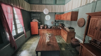 A vintage kitchen with muted green walls and checkered green flooring. The room features wooden cabinets, a long wooden table in the center, and an old-fashioned orange refrigerator. Various kitchen items, like pots and jars, are arranged on the cabinets. A round window and pink-patterned curtains add a retro charm.
