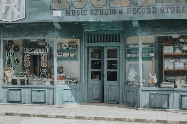 A vintage-style storefront with a teal facade, showcasing a variety of retro music equipment and devices in the windows. Large speakers, a reel-to-reel tape recorder, and other audio equipment are prominently displayed. A framed poster and a guitar add to the nostalgic ambiance. The shop sign indicates a music studio and record store.