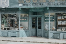 A vintage-style storefront with a teal facade, showcasing a variety of retro music equipment and devices in the windows. Large speakers, a reel-to-reel tape recorder, and other audio equipment are prominently displayed. A framed poster and a guitar add to the nostalgic ambiance. The shop sign indicates a music studio and record store.