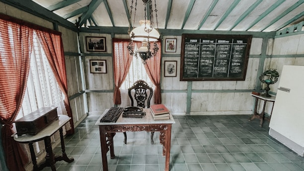 A vintage room features an intricately carved wooden desk at its center, topped with an old typewriter and books. Against the walls are framed photographs and a large chalkboard with writing. Red curtains with white lace filters light through the windows. The space is adorned with an ornate chandelier and a classic table lamp on a side table.