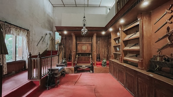 An interior room with antique wooden furniture and ornate decorations. The space features a red carpet and an intricately carved wooden throne at the center. Wall-mounted shelves display various antique items, including weapons like swords and spears. The lighting creates a dim, historic ambiance.
