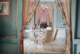 An interior scene featuring a cozy, retro-decorated room with green walls and patterned floors. The room includes a round table with a potted plant, surrounded by light-colored armchairs. Colorful, patterned curtains frame the entrance, adding to the vintage charm. A wooden cabinet is seen in the background, with additional decorative items.
