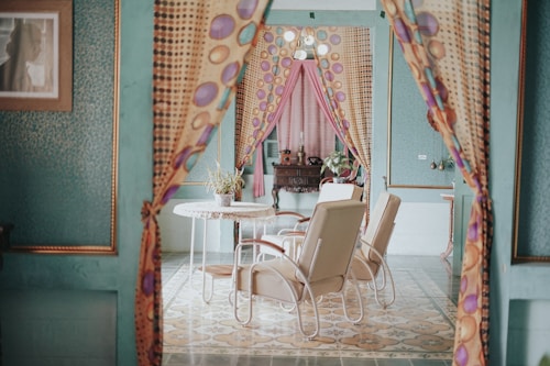 An interior scene featuring a cozy, retro-decorated room with green walls and patterned floors. The room includes a round table with a potted plant, surrounded by light-colored armchairs. Colorful, patterned curtains frame the entrance, adding to the vintage charm. A wooden cabinet is seen in the background, with additional decorative items.
