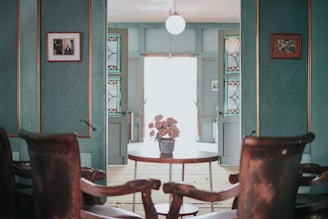 A vintage-style room with teal walls and wooden chairs set around a small round table. The table holds a decorative vase with flowers. The walls are adorned with framed photographs and stained glass panels. Ambient light from a globe ceiling lamp softly illuminates the space, lending it a cozy, nostalgic feel.