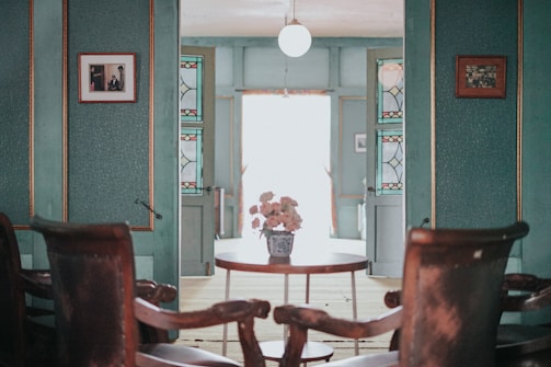 A vintage-style room with teal walls and wooden chairs set around a small round table. The table holds a decorative vase with flowers. The walls are adorned with framed photographs and stained glass panels. Ambient light from a globe ceiling lamp softly illuminates the space, lending it a cozy, nostalgic feel.